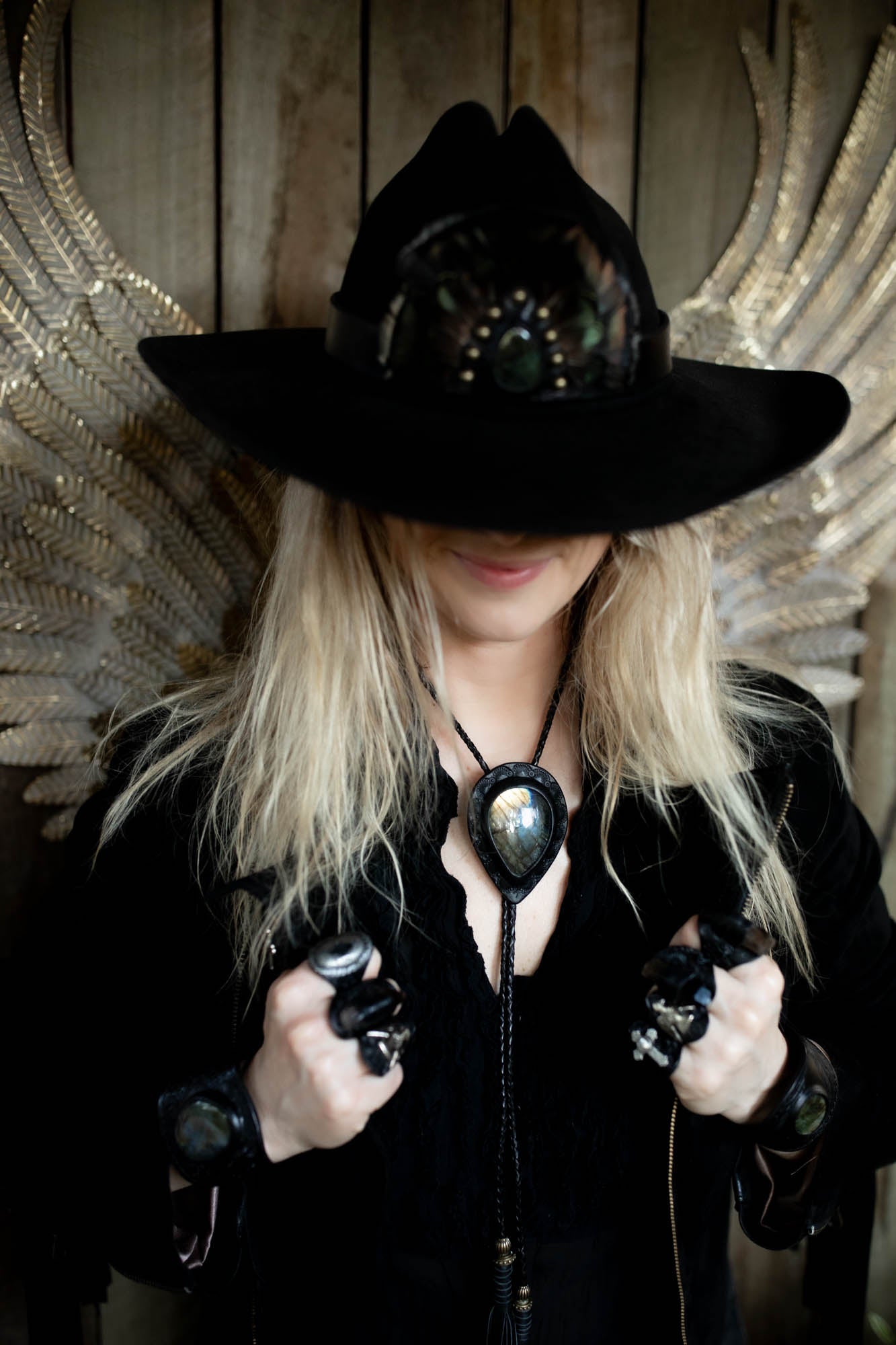 Person wearing a black hat and coat with decorative elements in front of a decorative wall with gold wings